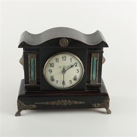 Chinese Wood Mantel Clock: A Korean wood mantel clock. The clock has a black finish and features a curved bonnet with a brass chrysanthemum crest. Other features of the clock include side columns with blue acrylic and brass accents and a hinged brass bezel and glass cover displaying a white face with black Arabic numbers and hands. The clock also displays decorative brass side handles with scrollwork trim to the base and the clock stands on ornate brass legs with trifid feet. The clock is labeled to the face in Chinese characters, reading “Registered trademark” (註册商標) with a character in a circle on he face reading “yong” (永).