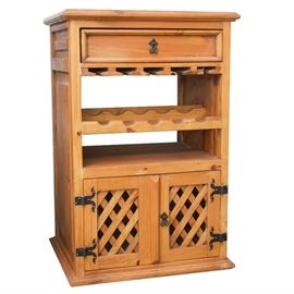 Wine Cabinet: A wine cabinet in pine. This cabinet features a rectangular top with a beveled edge over a drawer with dovetail joinery and decorative iron pull. The cabinet includes a wine glass storage rack, bottle rack, open shelf and a pair of lattice doors with decorative hinges and pull. The piece is unmarked and terminates in a plinth base. The piece has a two-sided design with access from both sides to drawer, shelving, and storage.