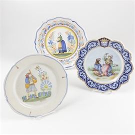 Henriot Quimper Faïence Plates: A selection of Henriot Quimper Faïence plates. The collection of hand-painted earthenware includes a plate featuring a central design of a Breton woman decorated with floral and leaf motifs and a scalloped edge; plate with a central design of a man playing bagpipes in a garden setting accented with blue rim and signed to the lower center; and a plate featuring a central design of a Breton man and women accented with a blue filigree border and yellow-gold trim along the scalloped edge, marked to the underside “Henriot Quimper, RB F.G.”.