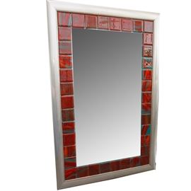 Red Tiled Wall Mirror: A wall mirror with red tiles. This mirror has a silver tone wood frame with red ceramic tiles and green grout that surround the edge of the mirror. The mirror has wire to the verso for hanging vertically.