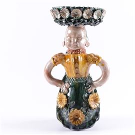 Mexican Earthenware Figural Pedestal Dish: A Mexican earthenware figural pedestal dish. The hand made dish is circular in shape and features floral decorations around the edge and balanced on the head of a female figure. She wears a yellow top with a floral green and yellow dress. There are no discernible maker’s marks.