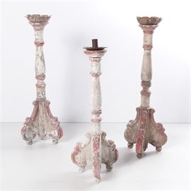 Large Floor Candlesticks: A group of large floor candlesticks. The three matching wooden items feature three ornate splayed legs which are supported by ball feet. Each turned column is topped by a metal sconce, with two of the sconces having an upward rising petal design. The smallest candlestick features a metal sconce with an egg and dart rim. The pieces are not marked with a maker or manufacturer name.