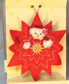 Steiff Teddy in a Red Star Ornament 714 in white  mohair.  'Shine on Teddy'.  This white teddy bear  with gold embroidered nose and mouth is within a  red felt star accented with gold thread.  Wears an  elephant ear button to mark the 100 year
