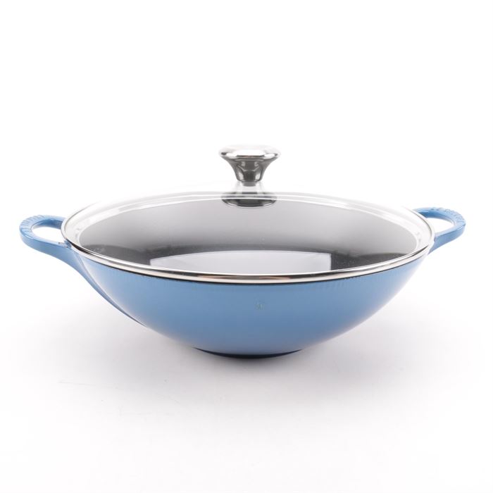 Le Creuset Enamel Wok: A Le Creuset enamel wok. This piece is presented in a blue finish with side handles and a removable glass lid. The lid is adorned with a metal frame and round knob that is embossed, “Le Creuset”. It is marked to the underside, “Le Creuset, 32, France”.