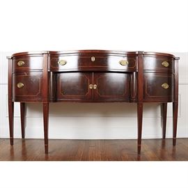 Vintage Hepplewhite Style Sideboard: A Hepplewhite style, mahogany sideboard, having a serpentine form. The front has a center drawer, slightly projected above two inlaid panel doors. Flanking each end, are a bowed drawer, over a drawer, each having inlaid borders and elliptical brasses. The case stands on six tapered-square legs with inlay and cuffed feet, to the forward four legs.