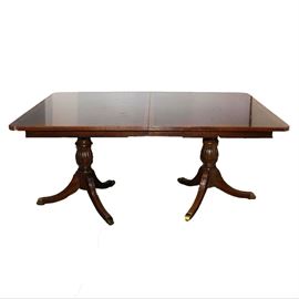 Federal Style Mahogany Dining Table with Double-Pedestal Base: A Federal style mahogany dining table with a double-pedestal base. The frame features a rectangular, crossbanded top with rounded corners and reeded edges above two turned standards. Each standard rises on three downswept, reeded legs ending in brass tone caps. There are no visible maker’s marks.