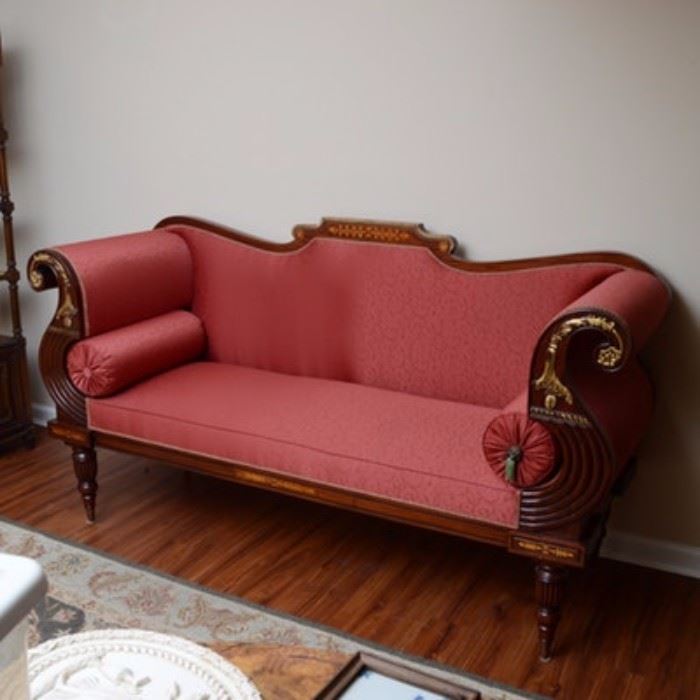 Closson's Empire Style Sofa: A Closson’s empire style sofa. This sofa is set on a wooden base and upholstered in a red vine pattern. The sofa features curved armrests fitted with cylindrical pillows with tassels on either end. The wooden frame features reeded details and a carved floral design. Some of these carved floral designs are painted in a gold-tone paint. The legs of this sofa are tapered. Located in lower level of home with access to garage.