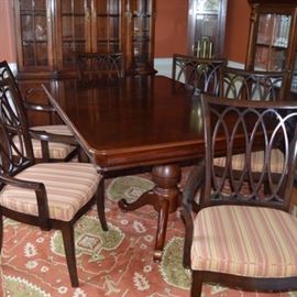 Dining Table and Eight Chairs: A dining table with eight armchairs. The wood table features a tabletop with veneer inlay in a diamond pattern and two large turned pedestal bases, each with four spider legs. The tabletop can be extended with the addition of two 15 inch leaves. It is accompanied by eight armchairs with arched back with openwork design of intersecting ovals. The armchairs include seats upholstered in a brick red and tan stripe fabric, which stand on square, tapered legs. The group is located on first level of home.