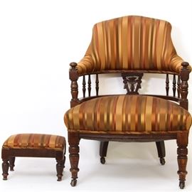 Upholstered Barrel Back Chair with Footstool: An upholstered barrel back chair with a coordinating footstool. The chair features a crested upholstered back rest over turned spindles and carved wood splat with turned stiles topped with knob finials. It is upholstered in a gold and orange toned striped fabric with turned front legs and saber rear legs including casters. A small rectangular wood framed footstool with a matching fabric top is also included.