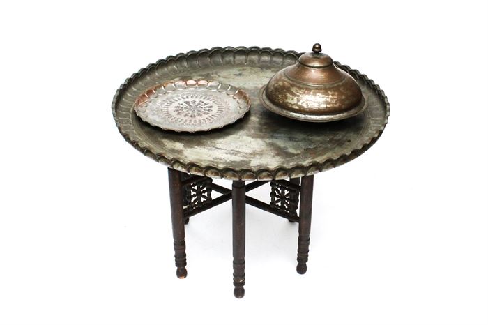 Antique Mid-Eastern Tinned Copper Tray Table, Tray, and Lidded Bowl: A selection of antique tinned copper ware. Included is a Mid-Eastern large tray table featuring a pie crust edge and a plain well. It is paired with a folding hand-crafted six-leg wooden base. A smaller tray features scalloped edges and an etched concentric design to the well. Both trays feature hanging hooks applied to the versos. A lidded bowl features a finial to the domed lid. All items are unmarked.