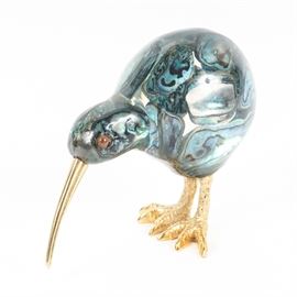 Art Glass Kiwi Bird with Gold Tone Beak and Feet: An art glass bird figurine. This piece depicts a kiwi bird with a blue, black, and green hued glass body with an iridescent finish resembling a Pāua shell. The bird features orange-hued glass eyes with a long gold tone metal beak and gold tone metal feet. The underside of one foot is embossed “0K”.