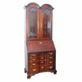 Bonnet Top Cherry Slant-Front Secretary: An American Colonial style secretary bookcase, having a bonnet top with double-arched and molded pediment, above two arched and framed, beveled glass doors, which open to a lighted interior with two glass shelves. Below is a slant-lid desk, which folds outward, to reveal a center prospect door, fluted pilaster drawers, two small drawers and six valanced pigeon holed. The base features a block-front, with three shaped drawers, each having pierced brass pulls and escutcheons, above carved bracket feet. Manufacturer unknown, circa 1980-90s.
