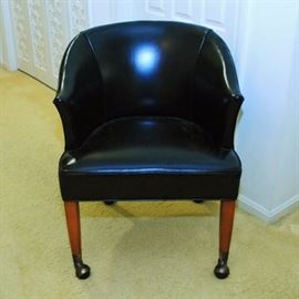 Black Leather Barrel Chair on Casters: A black leather barrel chair on casters. The black leather upholstery is trimmed in brass tacks along the back contour. The chair stands on four square tapered legs with brass caps and casters.