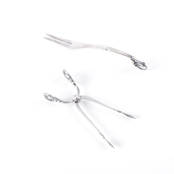 Georg Jensen "Ornamental" Sterling Fork and "Blossom" Sugar Tongs: A pairing of sterling silver flatware by Danish silversmith Georg Jensen. This includes a two-tine cold cut serving fork in the Ornamental #21 pattern, featuring a curved, Art Nouveau style handle with beaded and flower petal tip. It is stamped to the back “21 Denmark Sterling” alongside the “GI” beaded maker’s mark, dating this piece to circa 1910-1925. Also presented are a pair of sugar tongs in the Blossom pattern with leaf-shaped tips and flower bud designed handles. It is marked to one side “Sterling” and to the other “Denmark”. The total approximate weight is 1.335 ozt.