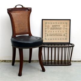 Vintage Chair, Needlepoint and Magazine Rack: A vintage chair, needlepoint and magazine rack. The chair features an arched crest rail with added brass-tone metal handle and a curved caned back rest. The round seat is upholstered in black faux leather with button tufting and nail head trim. The chair is tagged “Made by Hickory Chair Company, Hickory, North Carolina.” The walnut magazine holder is from “Cornwall Wood Products” and a framed sampler, wired to hang and with a personal inscription on the back.