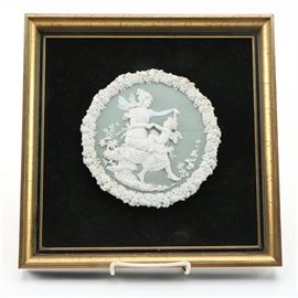 Low Relief Sculpture of Winged Figures: A low relief ceramic sculpture, rendered in blue and white in the style of Wedgwood porcelain, of winged figures. This piece depicts cupid on the lap of a fairy-winged woman, flowers and grasses at her feet. The piece is surrounded by a border of molded fruit and is presented against a black backing in a gold tone frame, a wire to the verso for hanging.