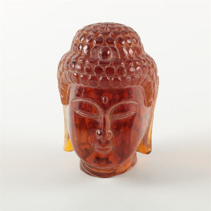 Amber Glass Buddha Head: An amber glass Buddha head sculpture. This piece depicts the long-eared figure with his eyes half-closed, and lips pursed. It does not have any visible maker’s marks.