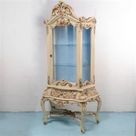 Louis XV Style Carved and Painted Curio Cabinet: An exuberantly carved and painted curio cabinet in the Louis XV style. An assymetrical scroll and acanthus leaf-carved pediment rests above a shaped glass door framed by floral and foliate carvings, flanked by matching sides with flat glass sidelights. The interior is lined in a blue plush and has two glass shelves. The separate base, features a carved drawer and apron, between four heavily carved cabriole legs and fanciful cross stretcher. Circa 1960s.