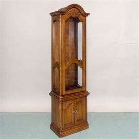 Oak Cabinet: An oak cabinet featuring an arched pediment over a hinged glass door. The arched door is embellished with an incised scroll based design, The sides are also glass paneled. The lower case features two hinged cabinet doors with brass-tone hardware. The doors have recessed panels and corners embellished with a floral and leaf design. The cabinet rests on a plinth base.