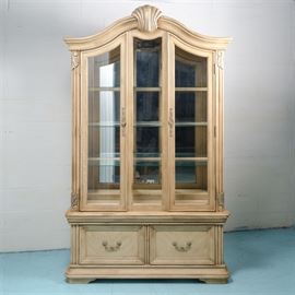 Contemporary Queen Anne Style China Cabinet: A Contemporary / Queen Anne style china cabinet. This two piece cabinet features a pickled oak stain and curved crest rail with molded trim and applied shell embellishment above two cabinet doors with glass panes opening to three glass shelves, a recessed light, and a mirrored back. The cabinet doors are flanked by fluted stiles with acanthus leaf detail. The lower base features two aligned drawers with recessed panels rising on bracket feet. The back is marked on a sticker.