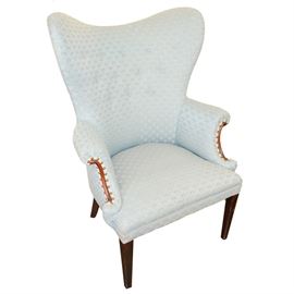 Vintage Upholstered Wing Back Chair: A vintage upholstered wing back chair with a dramatic curved back and wings, rolled arms and seat cushion. The arms feature a scrolled wood trim surrounded by brass tacks and gimp. The chair stands on two square tapered front legs and two slightly splayed back legs. The upholstery fabric is pale blue with a textured fan pattern.