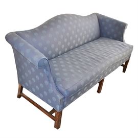 Upholstered Camel Back Sofa: A camel back upholstered sofa with rolled arms and a single seat cushion The sofa is upholstered in a blue fabric with a shell print. The piece rests on a wooden frame with six square legs connected by an H stretcher. The front legs feature low relief carvings.