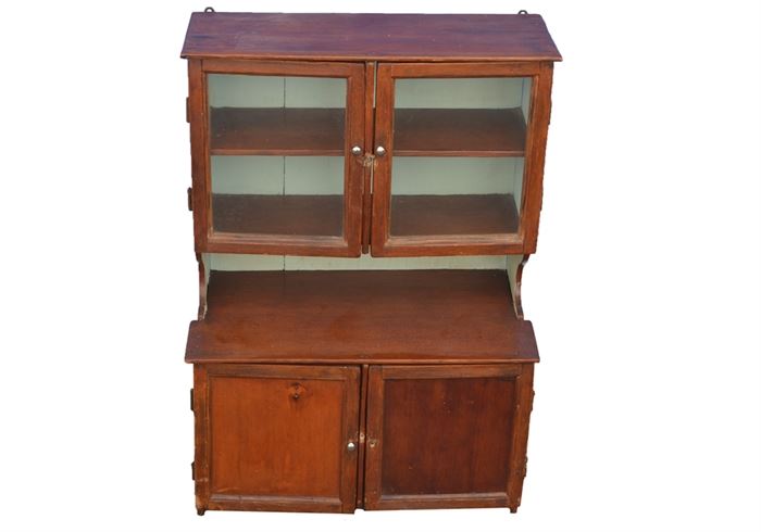 Cabinet with Display Hutch: A vintage cabinet with display hutch. This cherry stained wooden piece has a simple cabinet at the top with two wooden shelves behind glass paned doors, set over a workspace with curved side panels. The work space is the top of a lower cabinet, which has two wooden shelves behind blind panel doors. The doors all have metal knobs and the top has a metal latch. The back is made of long, vertical boards, painted a creamy tan.