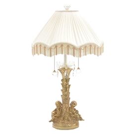 Cast Brass Cherub Table Lamp: A cast brass cherub table lamp. The lamp features two light sockets with faceted glass pull chains, above a scalloped glass bobeché with attached faceted glass prisms. The cast brass base depicts two cherubs seated at the base of a tree trunk, being topped with a cream fabric covered shade with fringed accents and a faceted glass finial. The lamp is unmarked by the manufacturer.