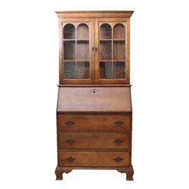 Colonial Style Drop Front Secretary: A Colonial style drop front secretary. This rectangular top secretary with walnut stain and molded trim features two cabinet doors with arched glass panels and mullions opening to two shelves. The drop front panel features a batwing escutcheon. The lower base features three full width drawers with brass batwing plates and bail handles terminating on ogee bracket feet. There are no visible maker’s marks.