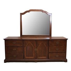 Chest of Drawers with Mirror by Century: A wooden chest of drawers and a mirror by Century Furniture. This horizontal dresser features central cabinet doors holding three drawers with teardrop pulls flanked by three machine dovetailed drawers with batwing pulls on either side. It stands on ogee bracket feet between a carved apron. A manufacturer stamp is seen to the interior of the drawer. The rectangular wood framed beveled mirror includes a scalloped arched top.