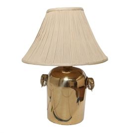 Brass Tone Table Lamp: A brass tone table lamp. The base features a bulbous shape and is composed of metal with a brass tone finish. The sides of the lamp are each decorated with elephant style knobs and the lamp features an off white empire style lamp shade.