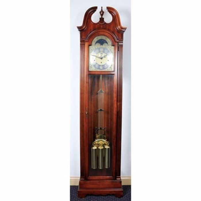Howard Miller Grandfather Clock: A Howard Miller grandfather clock. The clock is hardwood and veneers with a cherry finish and features a broken pediment bonnet with an urn-shaped finial and reeded side columns. Other features include wood and glass side panels and a glass front door that display a silver-tone face with raised brass numbers and black serpentine hands surrounded by decorative brass floral and scrollwork spandrels. Other features include a moon phase dial displaying the stars on a night sky, a glass front center door with a brass lock and key that displays a brass pendulum and weights and the clock stands on a plinth style base with bracket feet. The clock has a key and is labeleled “Howard Miller” to the face.