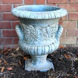 Concrete Urn Planter: An urn shaped concrete planter. The planter has grapes embossed around the body of the urn. There is no maker’s mark visible on the planter.