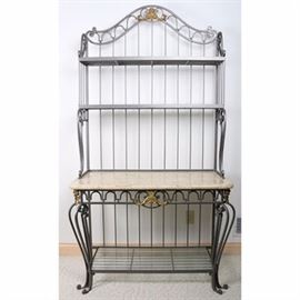 Metal and Marble Top Baker's Rack: A metal baker’s rack. It has a dark grey metal frame with arched top, scrolled sides, and vertical rods along the sides and back. It has two glass shelves recessed above a tan marble top, which sits on an elaborate apron and cabriole legs, over a third glass shelf. The pediment and apron have gold colored accents.
