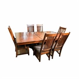 Arts-and-Crafts Style Dining Table and Six Chairs: An Arts-and-Crafts style dining table and six chairs. The table features an oak top with one leaf, having butterfly or bowtie inlay. The top is on a square-spindled trestle base with shelf stretcher. The matching chairs consist of two arm and four side chairs in stained alder, each having a tall square spindled-back, over an upholstered seat, which is covered in a blue foliate pattern. The chair bases feature spindled sides and an arched seat apron.