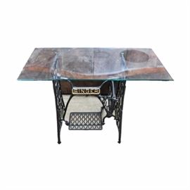 Glass Top with Vintage Singer Wrought Iron Sewing Machine Base: A glass top with Singer wrought iron base. The table features a rectangular sheet of tempered glass over a vintage Singer wrought iron sewing base . The base is a treadle model with gold painted accents.