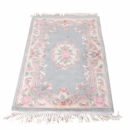 Chinese Aubusson Style Area Rug: A Chinese Aubusson style area rug. This wool area rug features a color palette of pastel pink, green, blue and cream. It features a scalloped pink rose medallion on a pale blue field with a scroll border and pink tonal floral brackets at the corners and sides. It has white colored applied fringe to either end and it is marked “Made in China”.