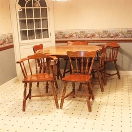 Vintage Traditional Style Dining Table and Chairs: A vintage traditional style wooden dining table and six chairs. The round extendable table features a beveled edge and stands on four turned and splayed legs. Four side chairs and two barrel back arm chairs each comprise a scalloped crest rail over a spindle back and carved seat. They rise on four turned, splayed legs with turned H-style stretchers. No maker’s mark present. Two leaves are also included.