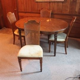 Vintage Broyhill Federal Style Dining Table and Chairs: A vintage Federal style dining table with chairs by Broyhill. This set includes an oval top table with oak stain and beveled edge overhanging thin apron sides rising on tapered legs. Also includes four dining chairs with curved crest rail and wicker back panels flanked by finial topped stiles. The seats are upholstered in an off white fabric and rises on tapered legs. The chairs are marked “Broyhill” to the underside. One table leaf is included.