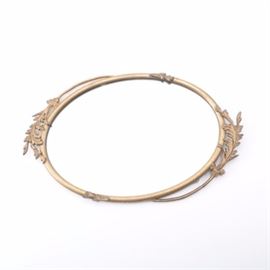 Metal Leaf Themed Mirror: A metal leaf themed mirror. The mirror is oval in shape and features a gold tone metal frame. The frame is decorated on either end with lancet leaf style accents and the mirror features a wire hanger to the back.