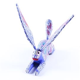 Wood Carved Stylized Rabbit Figurine: A wood carved stylized rabbit figurine. The figurine depicts a rabbit laying on the ground with an elongated tail and large ears, all painted in blue with patterns in white, red, and light green. The rabbit is marked “Pepe Santiago” to the underside and the ears are removable.