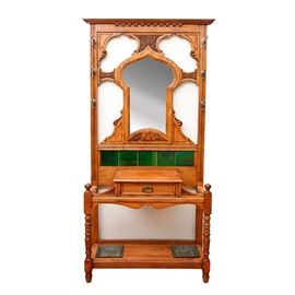 Antique English Carved Oak Hall Tree: An antique English carved oak hall tree. This rectilinear hall tree features a mirror in the shape of an Islamic arched window joined on four sides by carved panels which connect to the frame of the hall tree. Above the mirror is a strip of dental carving just below the cornice; on each side of the frame are three up turned silvered metal hooks which end in acorn finials. Below the mirror and enclosed by an oak frame are five glossy dark green ceramic tiles which sit above a protruding storage box with a single drawer for gloves which is secured in the front upon a horizontal support, with carved spandrels above and a decorative scroll below, between the two turned front legs of the hall tree. In the two open areas on either side of the storage box and between the outer frame are placed the umbrellas whose tips rest on the metal drip pans set into bottom shelf of the hall tree which is directly above the turned blunt arrow feet. Unmarked.