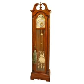 Sligh Grandmother Clock: A Sligh grandmother clock. The clock has a cherry wood and veneer case with a broken pediment and gold-tone finial on an arched bonnet with reeded side columns that have ring turned wood caps. The clock has a glass front door that displays a silver-tone metal face with raised gold-tone Arabic numbers and serpentine hands. The face has a gold-tone metal backdrop with spandrels, a small second clock and a brass moon phase clock above. Other features of the clock include a lyre style pendulum and weights with a gold-tone finish and the clock has a curved apron and stands on bracket feet with a tassel accent to the door. The clock has a key for winding and an instruction manual. The clock is labeled to the base “Slight” and is untested.