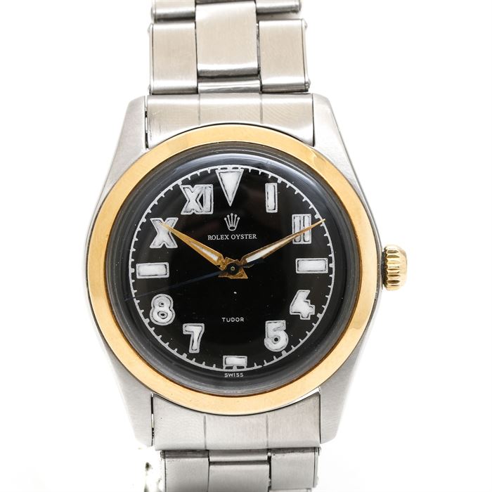 Rolex Oyster Stainless Steel Wristwatch: A Rolex Oyster stainless steel wristwatch. This wristwatch features a Rolex rivet band. The case has a yellow gold plate bezel. A black dial displays alternating Arabic numeral and Roman numeral hour markers, accented by luminous styled hands.