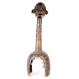 West African Metal "Vertical Mask": A metal sculpture from Nigeria, west Africa, in the manner of Mumuye and Wurkun tribe vertical masks. The sculpture is made from solid brass or bronze, and depicts the head of a man atop a round column with raised circle designs, resting on an arched base, also with raised circle patterns. The head has exaggerated facial features, with an elaborate hairdo, holes through the ears, open eyes, and an open mouth with three-dimensional teeth. This piece appears to be created to place on a tall shaft or pole to display prominently, and is unmarked.