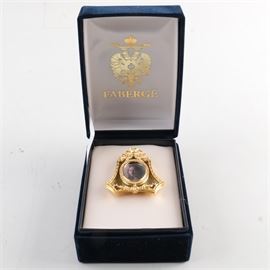Faberge Gold Tone Metal Miniature Frame: A Faberge gold tone metal miniature frame. The piece is oval in shape and features a ribbon style finial and a gold tone metal garland style accent along the base. It is decorated with colored and clear stones throughout and is marked to the back “Faberge”. It is accompanied by a box.