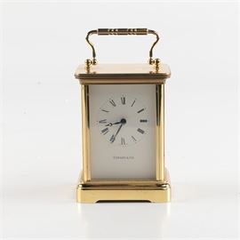 Tiffany & Co. Carriage Clock: A carriage clock by Tiffany & Co. This battery-operated clock is housed in a rectangular gold-tone metal case, with a simple square pedestal and handle at the top. Its dial features Roman numerals. The clock has not been tested, and time-keeping accuracy is not guaranteed.