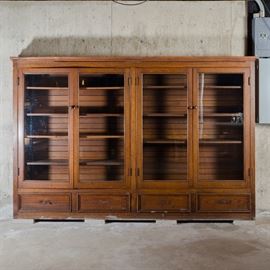 Oak Cupboard with Drawers: An oak cupboard with drawers featuring a rectangular top over two sets of glass-paneled doors that open to wood shelving and a horizontal slat back. The bottom is fitted with four drawers with recessed panels. Note, the back panels extend beyond the frame in back.