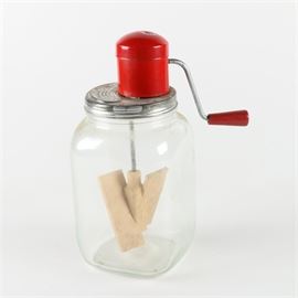 Hand Crank Mixing Jar: A hand crank mixing jar. This clear glass mixing jar features a metal lid with a hand crank mixer with wooden mixing paddles attached. The jar is unmarked.