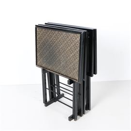 Folding Tray Table Set by Artex: A vintage set of four tray tables in a stand from Artex. Each table comprises a rectangular top with raised edges and includes a gold and black patterned top. They stand on foldable black painted “A” frame legs. A black stand is also included.